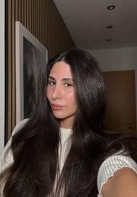 Portrait d'une personne aux cheveux longs et foncés, portant un pull blanc texturé, devant un mur rayé doucement éclairé avec une porte en bois.