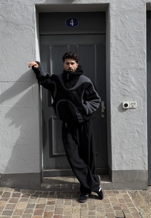 Vyras juodos ir pilkos fliso striukės bei juodų kelnių, atsirėmęs į pilkas duris su skaičiumi 4, stovintis akmeniniu grindiniu.