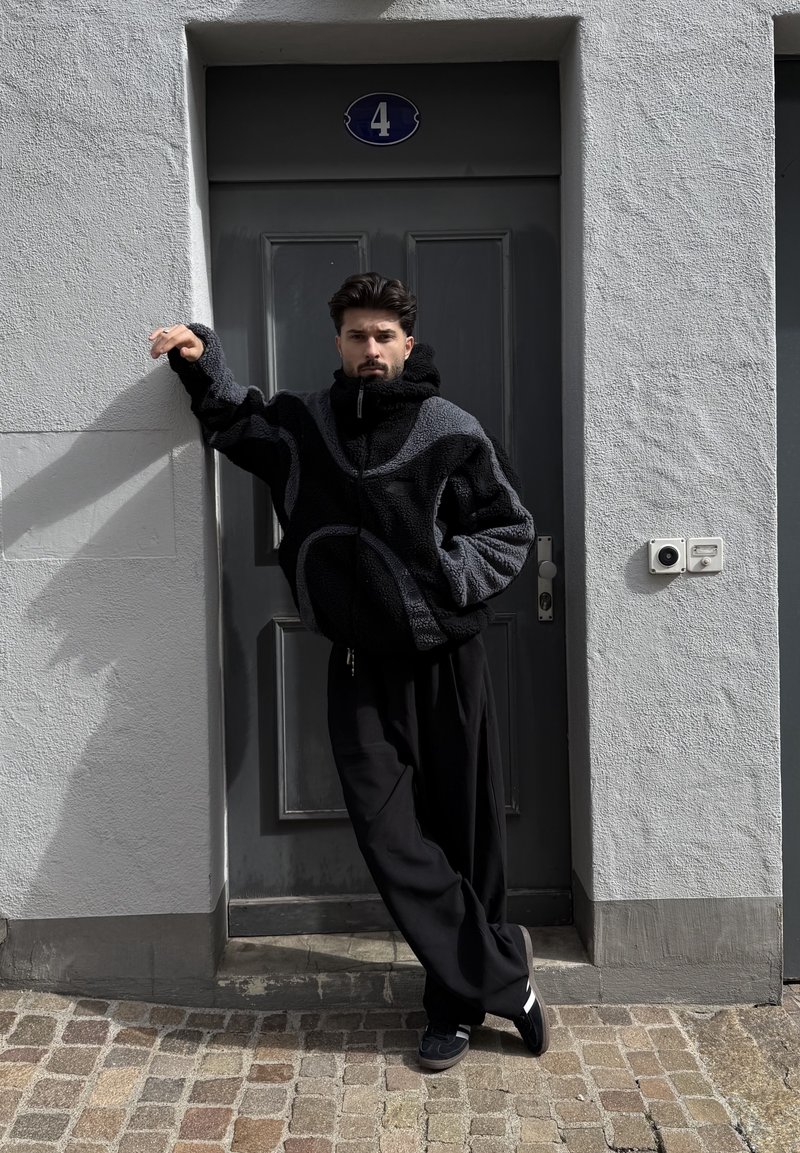Homme en veste polaire noire et grise et pantalon noir appuyé contre une porte grise numérotée 4, debout sur un pavé.