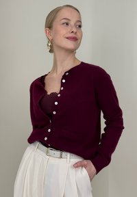 Cardigan bordeaux avec un col rond, fermeture à boutons, et petite poche avant, associé à une ceinture blanche et un pantalon plissé crème.