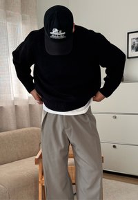 Pull noir en maille côtelée sur une chemise blanche, associé à un pantalon gris ample. Une casquette noire avec broderie blanche repose sur la tête. Fond neutre.