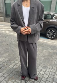 Persoană purtând un costum gri supradimensionat, maiou alb cu dungi, mai multe inele argintii, un colier cu cruce și pantofi loafer burgundy pe o stradă pietruită.