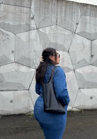 Macacão de ganga azul, com um design ajustado, acompanhado por uma mala tote de couro preta. Cabelos longos e escuros presos com um clip, encostada a uma parede de betão texturizada.