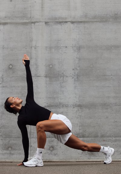 Top negro de manga larga, pantalones cortos blancos y zapatillas blancas. El sujeto está en movimiento, con un brazo levantado, posicionado contra una pared de concreto.