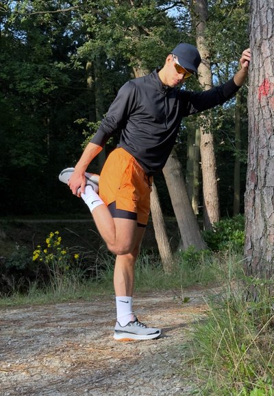 Hombre atlético con una camiseta negra de manga larga estira una pierna vistiendo pantalones cortos naranjas y zapatillas de correr grises con acentos naranjas, al aire libre.