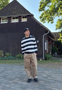 Suéter a rayas azul marino y blanco, pantalones beige holgados, zapatos negros y una gorra negra. Entorno al aire libre con un edificio de madera y vegetación.