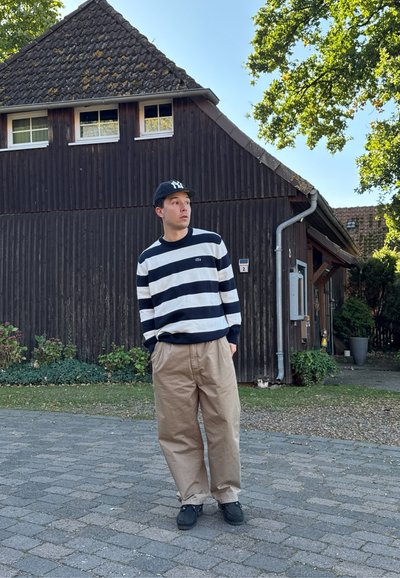Suéter a rayas azul marino y blanco, pantalones beige holgados, zapatos negros y una gorra negra. Entorno al aire libre con un edificio de madera y vegetación.