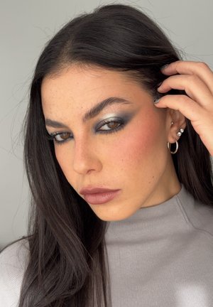 Primer plano de una persona con el cabello largo y oscuro, vistiendo ropa gris acanalada, con maquillaje de ojos ahumados y pendientes de aro plateados.