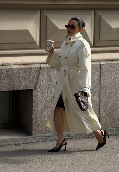 Mujer vestida con un largo abrigo trench color crema, tacones slingback negros y gafas de sol, llevando un bolso negro tejido y sosteniendo una taza de café mientras camina.