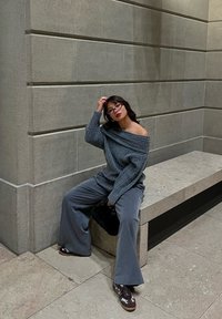 Femme portant des lunettes, habillée d'un pull gris à épaules dénudées et d'un pantalon ample, assise sur un banc en pierre devant un mur en pierre texturé, tenant un sac noir.