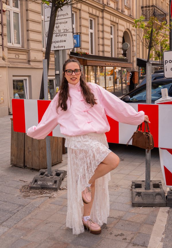 Junge Frau mit Brille, pinker Jacke und weissem Spitzenrock posiert auf einem Bein neben einer rot-weissen Strassenbausperre.