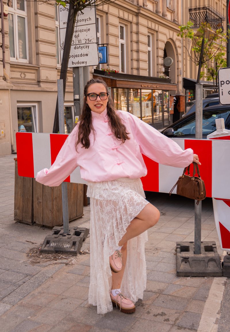 Noor naine, kes kannab prille, roosat jopet ja valget pitsseelikut, poseerib ühel jalal punase-valge tänavaehituse takistuse kõrval.