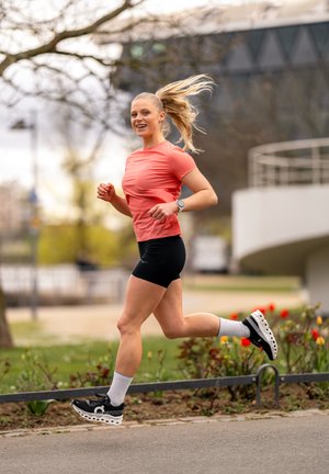Joven mujer haciendo jogging al aire libre cerca de un macizo de flores, vistiendo una camiseta coral, pantalones cortos negros, calcetines blancos y zapatillas negras de correr, sonriendo con la coleta al viento.