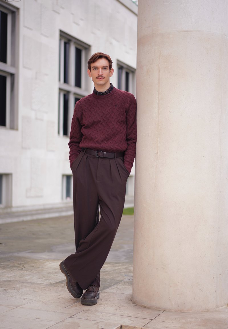 Jeune homme avec moustache portant un pull bordeaux et un pantalon marron, appuyé contre une grande colonne blanche à l'extérieur d'un bâtiment avec des fenêtres.