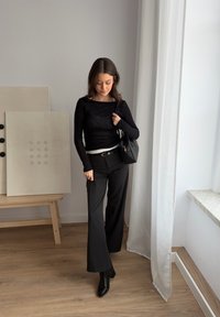Femme en haut et pantalon noirs à manches longues, debout à l'intérieur près d'un rideau blanc, tenant un sac à main noir, avec une œuvre d'art minimaliste sur un chevalet en bois derrière elle.