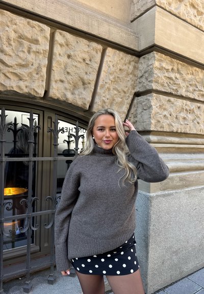 Mujer con cabello largo y rubio, vestida con un suéter de cuello alto gris y una falda negra con lunares, de pie cerca de un edificio de piedra con una ventana con rejas.