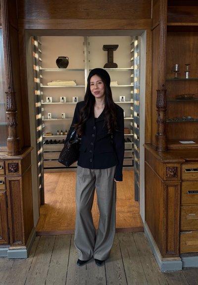 Joven mujer que lleva un blazer negro, boina y pantalones anchos grises, de pie dentro de una puerta con marco de madera con estantes y decoración detrás de ella.