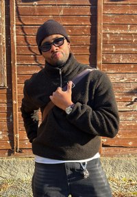 Homme portant un bonnet noir, des lunettes de soleil et une veste polaire, tenant une sangle marron sur l'épaule, debout devant un mur en bois vieilli.