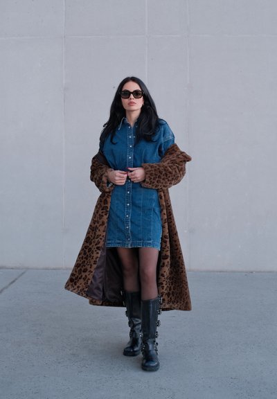 Mujer con gafas de sol, vestido de mezclilla, abrigo largo de estampado de leopardo, medias negras y botas negras con hebilla, caminando sobre concreto frente a una pared gris.
