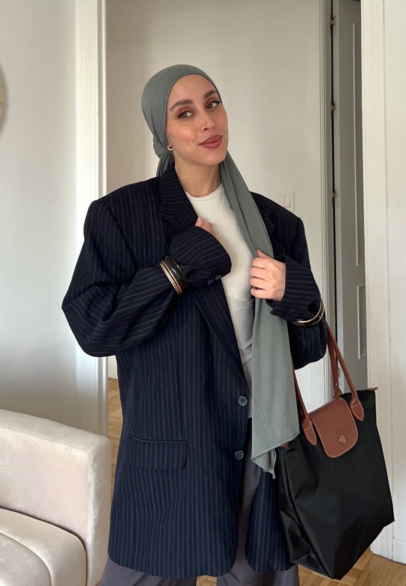 Femme portant un hijab gris, un blazer bleu marine à fines rayures oversize, une chemise blanche, tenant un sac cabas noir et marron à l'intérieur près d'un mur blanc.