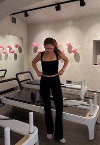 Sort atletisk top og bukser på en person i et fitnessstudie med Pilates-reformere og lyserøde dekorative kugler på væggen.