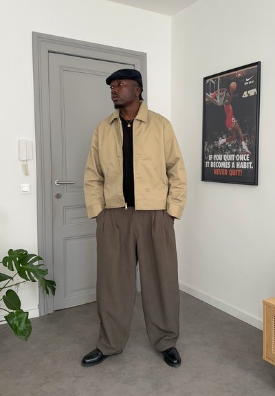 Hombre con chaqueta beige, camisa negra, pantalones anchos marrones y gorra negra, de pie en una habitación con puerta gris y cartel motivacional de baloncesto.