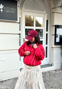 Rød bomberjakke med hvide kanter, lag på en grå top, kombineret med et beige, lagdelt, skinnende skørt og en mørkerød kasket med tekst.
