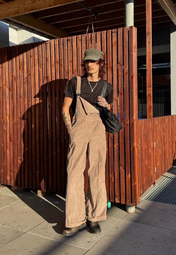 Junge Person mit Tätowierungen trägt beige Latzhose, schwarzes T-Shirt, graue Kappe und schwarze Stiefel und steht bei einem hohen Holzzaun im Sonnenlicht.