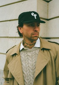 Svart keps med vitt broderat logotyp, beige trenchcoat och grå kabelstickad tröja. Visbara tatueringar och en örhänge. Neutral bakgrund.