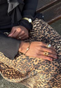 Mains ornées d'anneaux en or, bracelet en or et montre en or reposant sur une jupe à imprimé léopard, portant un haut à manches longues noir, assise sur un banc en bois.