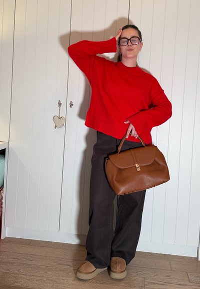 Mujer con suéter rojo y pantalones negros sosteniendo un gran bolso marrón, de pie con una mano en la cabeza y los ojos cerrados en interiores.