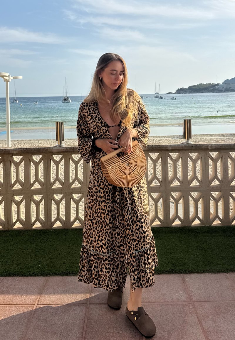 Femme en robe à imprimé léopard tenant un sac en bois en forme d'éventail, debout près d'une rambarde en bord de mer avec des voiliers en arrière-plan.