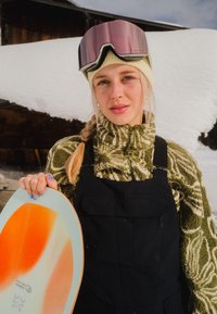 Jeune femme en tenue de ski avec des lunettes de protection roses tient un snowboard au design orange et blanc, debout dans un décor extérieur enneigé.