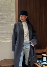 Femme portant un long manteau gris, un chapeau noir, un col roulé blanc et un jean bleu clair, tenant un sac à main noir, debout près d'un meuble en bois.