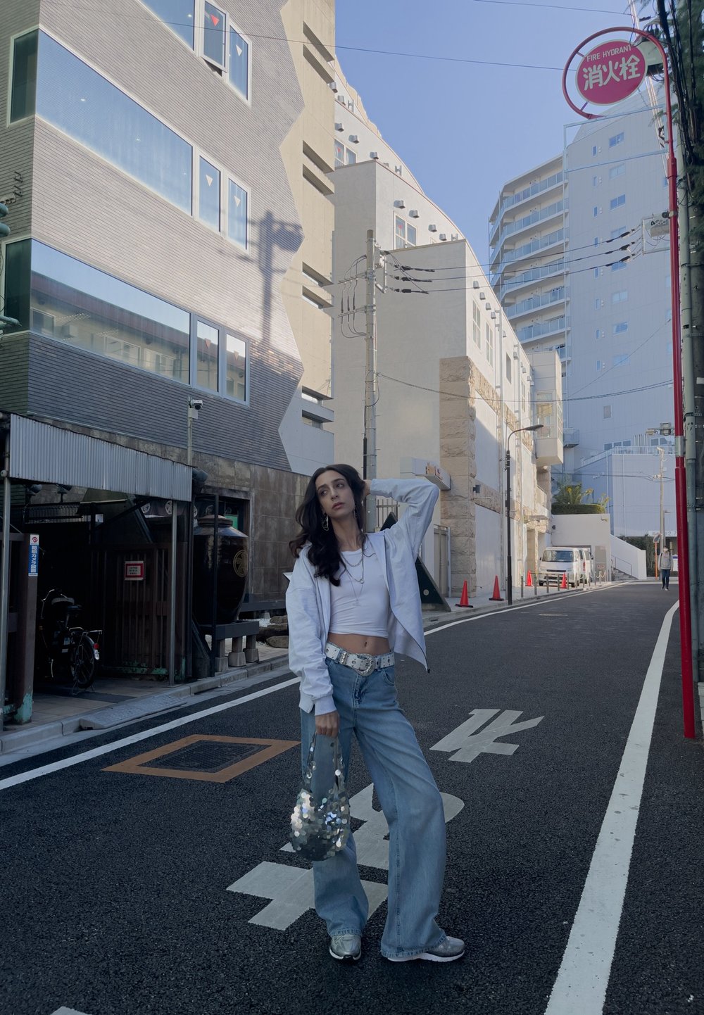 Καθημερινό σύνολο που περιλαμβάνει ένα λευκό κοντό μπλουζάκι, ανοιχτό μπλέ φαρδιά τζιν και μια ασημένια τσάντα με στρας. Το φόντο περιλαμβάνει αστικά κτίρια.