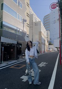 Καθημερινό σύνολο που περιλαμβάνει ένα λευκό κοντό μπλουζάκι, ανοιχτό μπλέ φαρδιά τζιν και μια ασημένια τσάντα με στρας. Το φόντο περιλαμβάνει αστικά κτίρια.