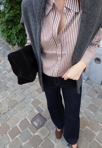 Rjava in bela prugasta srajca s gumbi pod sivoj cardiganom, v kombinaciji z temnimi zvonastimi kavbojkami in rjavimi loafersi, drži črno suknjič iz semiša.