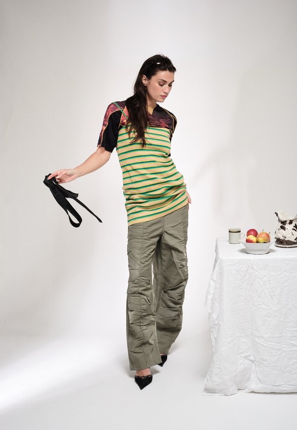 Γυναίκα με ριγέ πουκάμισο και χακί παντελόνι κρατάει μαύρα λουριά, στέκεται δίπλα σε ένα τραπέζι με ένα μπολ με μήλα, μια κούπα και μια κεραμική κανάτα.