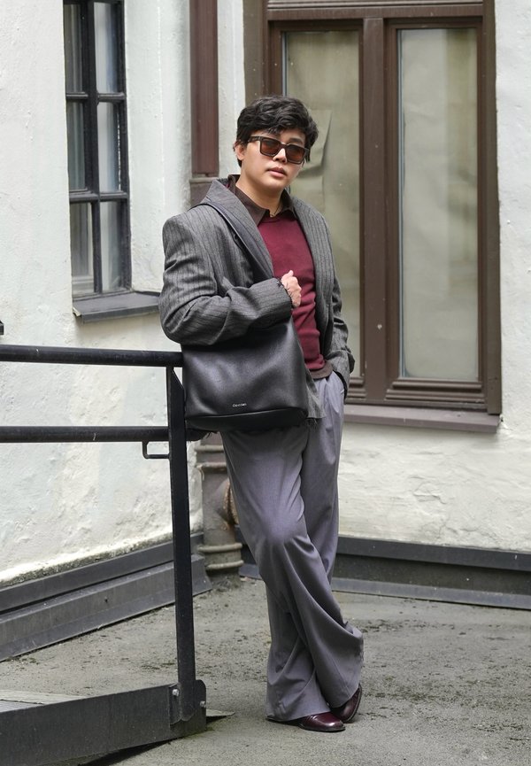 Persona que lleva gafas de sol, blazer gris, suéter burdeos, pantalones grises y zapatos oscuros, apoyada en una barandilla, sosteniendo un bolso negro al aire libre.