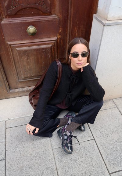 Cardigan negro de punto con cierre de lazo, top ciruela, pantalones de satén negros, calcetines gris-marrón y zapatillas New Balance negras con detalles en morado.