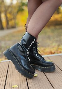 Schwarze Lederstiefeletten zeichnen sich durch einen hohen Glanz, genietete Ösen, eine dicke Gummisohle mit strukturiertem Griff und einen gelben Nähakzent aus.