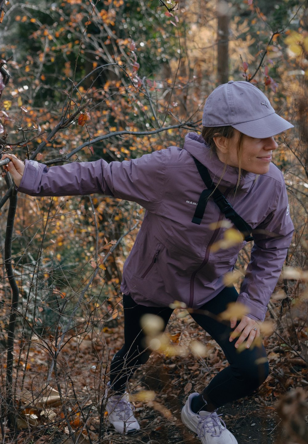 Giacca impermeabile viola chiaro con tasche con zip, leggings neri e sneakers bianche, indossata da una persona che si muove tra il fogliame autunnale.