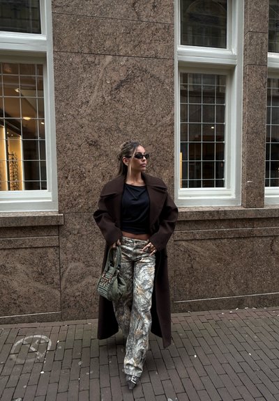 Mujer con abrigo oscuro, camiseta negra, pantalones estampados, gafas de sol y bolso verde, de pie junto a una pared de piedra marrón con ventanas.