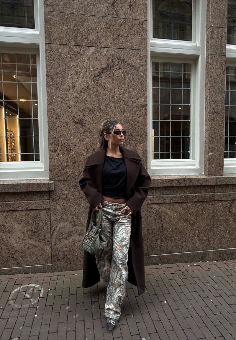 Γυναίκα με σκούρο παλτό, μαύρο τοπ, παντελόνι με σχέδια, γυαλιά ηλίου και κρατώντας πράσινη τσάντα στέκεται μπροστά σε καφέ πέτρινο τοίχο με παράθυρα.