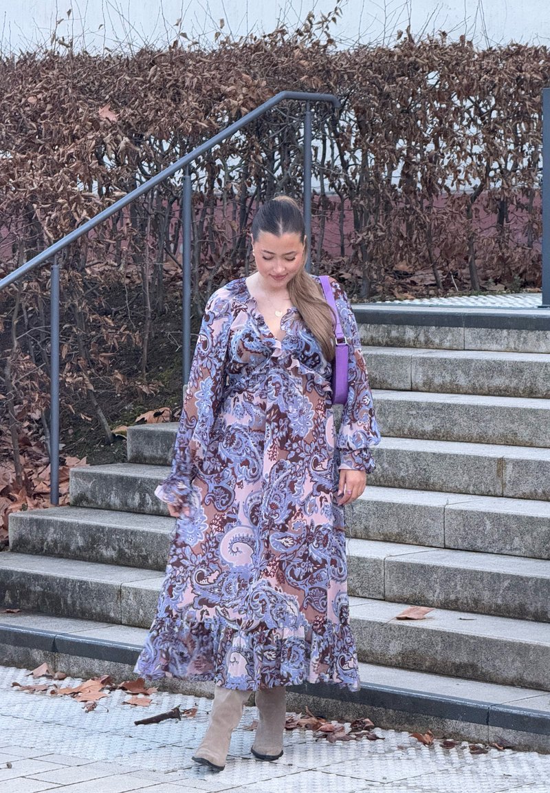 Vestido longo e fluido com um padrão paisley, apresentando tons de púrpura e castanho. Tem mangas longas e uma bainha com volantes. Botas bege até ao joelho.