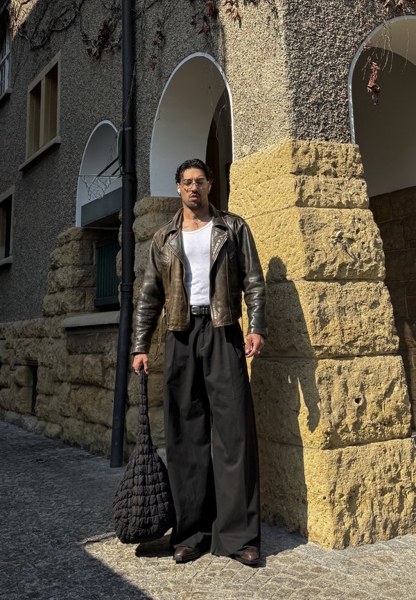 Homme portant une veste en cuir marron, un débardeur blanc, un pantalon noir large, tenant un grand sac noir texturé, debout près d'un bâtiment en pierre avec des arches.