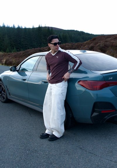 Hombre con gafas de sol, camisa de manga larga color granate, pantalones anchos blancos, apoyado en un BMW i4 azul estacionado en la carretera cerca del bosque.