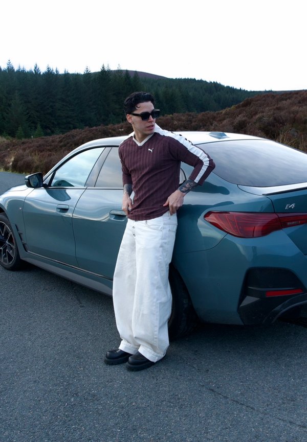 Uomo che indossa occhiali da sole, camicia bordeaux a maniche lunghe, pantaloni bianchi ampi, appoggiato a una BMW i4 blu parcheggiata su una strada vicino alla foresta.