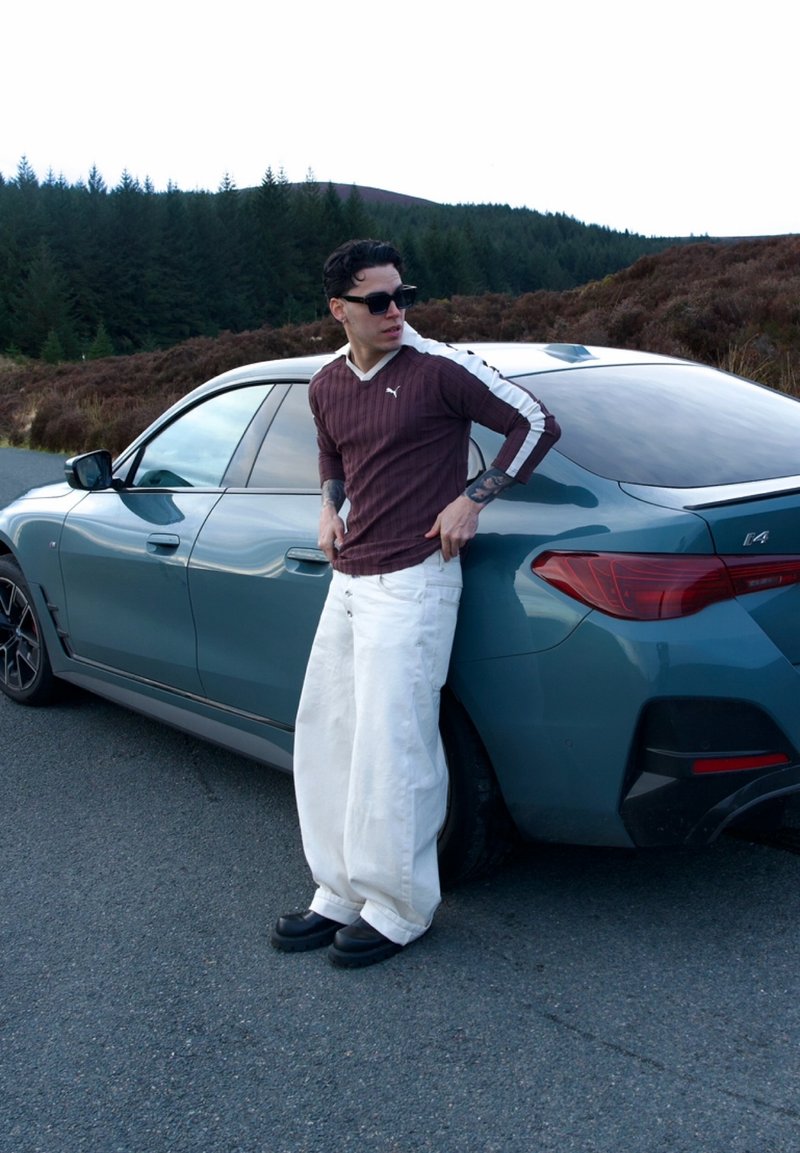 Man draagt een zonnebril, een bordeauxrode lange mouwen shirt en witte wijde broek, leunend tegen een blauwe BMW i4 geparkeerd op de weg nabij het bos.