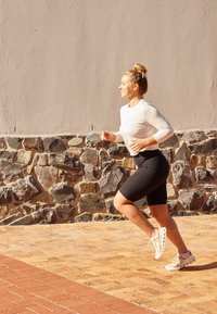 Sportoutfit mit einem leichten Langarmoberteil und schwarzen Shorts. Der Läufer trägt beiges Schuhwerk mit orangefarbenen Akzenten und bewegt sich auf einem Kopfsteinpflasterweg.
